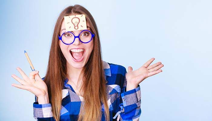 Smiling woman with glasses and a sticky note lightbulb on her forehead, holding a pen.