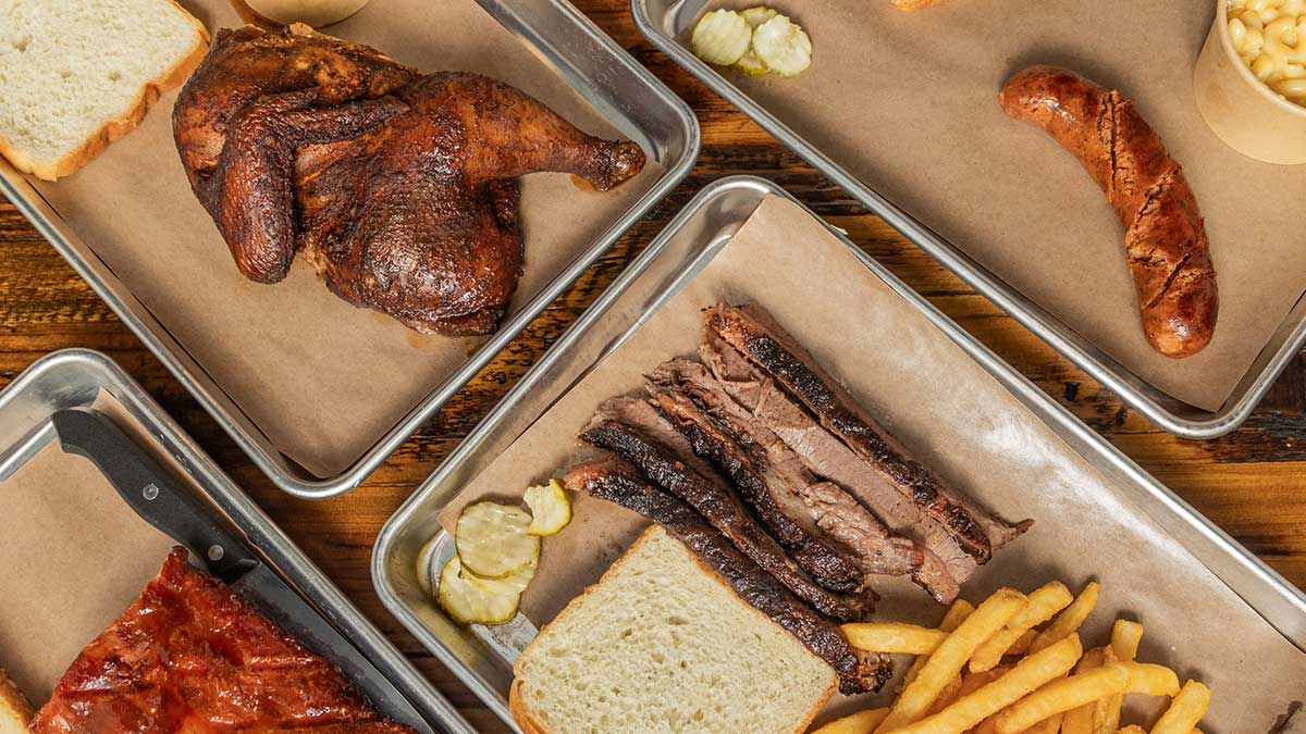 Assorted barbecue meats, fries, bread, pickles, and mac and cheese served on metal trays.