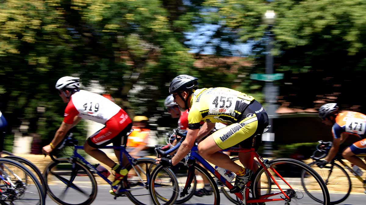 Cyclists in numbered jerseys racing on a road with trees and spectators in the background.