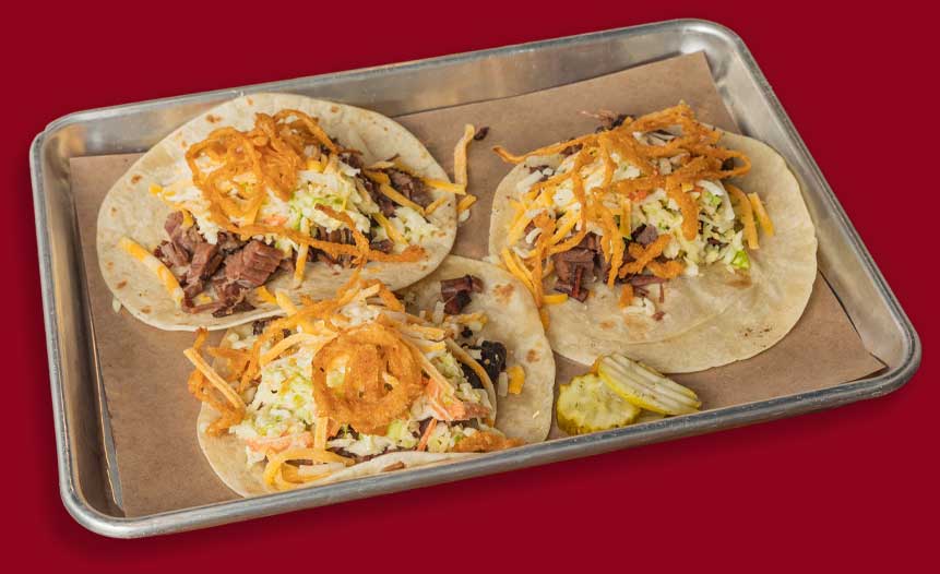 Three beef tacos with shredded cheese, slaw, and crispy onions at a BBQ restaurant in Mundelein IL.