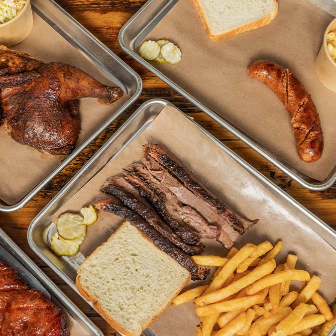 Three trays at a BBQ Restaurant in Mundelein IL serve meats, bread, fries, pickles, and sausage.