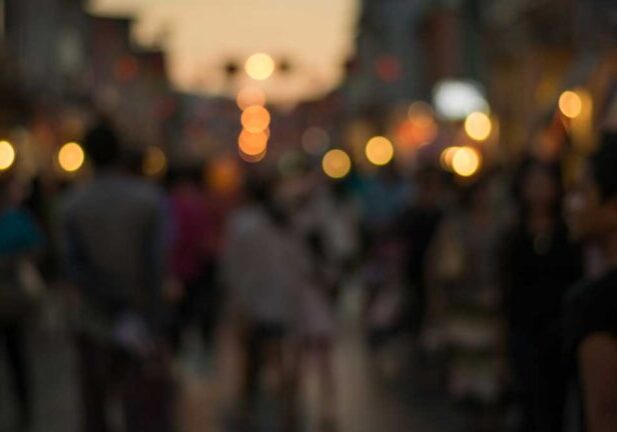 A blurred crowd on a street at dusk with warm, glowing lights in the background.